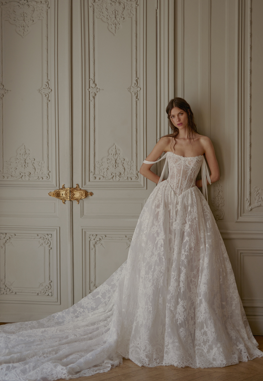 A woman in a strapless, lace wedding gown with a long train stands in front of ornate, cream-colored paneled doors.