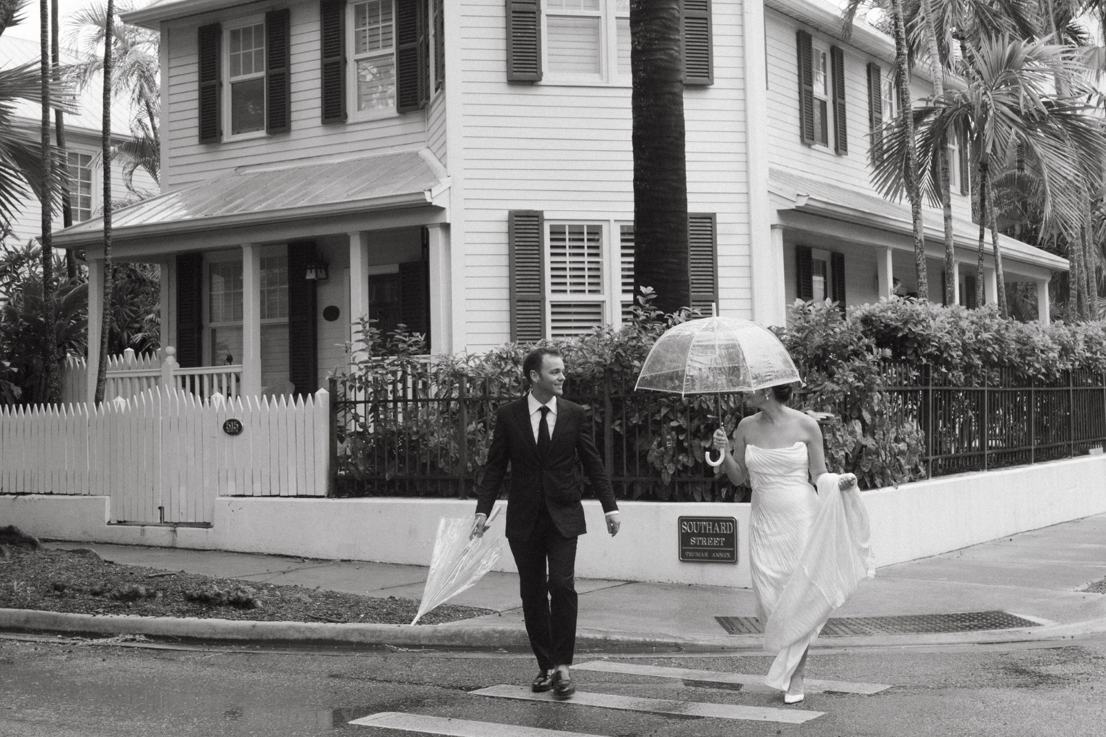 A man in a suit and a woman in a wedding dress cross a street holding transparent umbrellas in front of the Hemingway House on a rainy day in Key West, Florida.