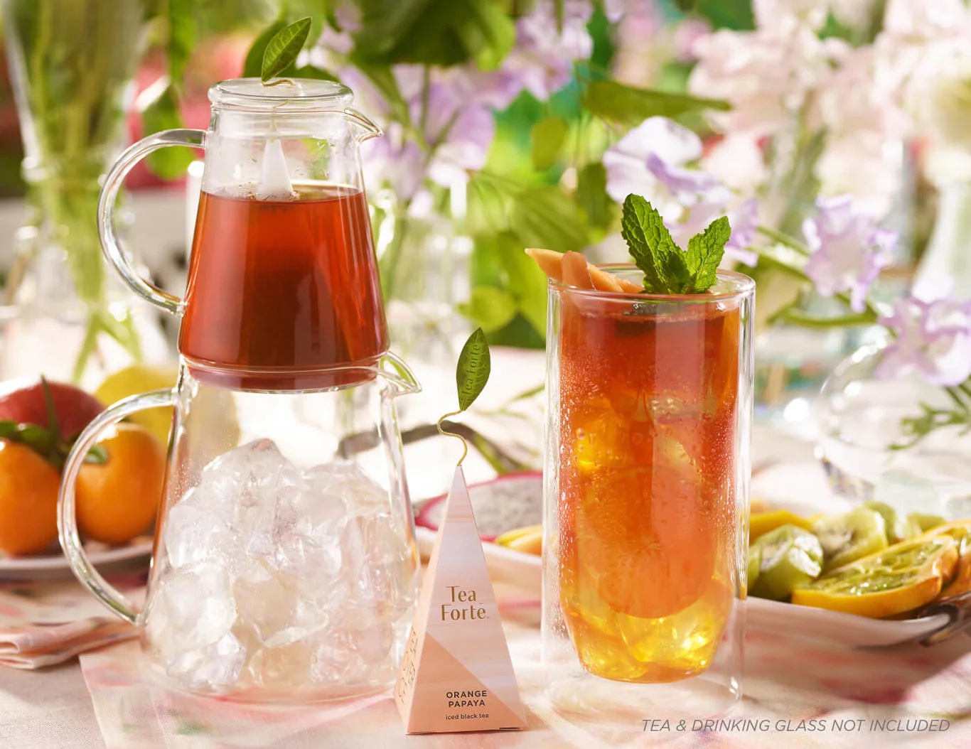 A glass pitcher with iced tea, a tall glass of iced tea garnished with mint and citrus, a tea bag, and assorted fruits on a floral-decorated table.