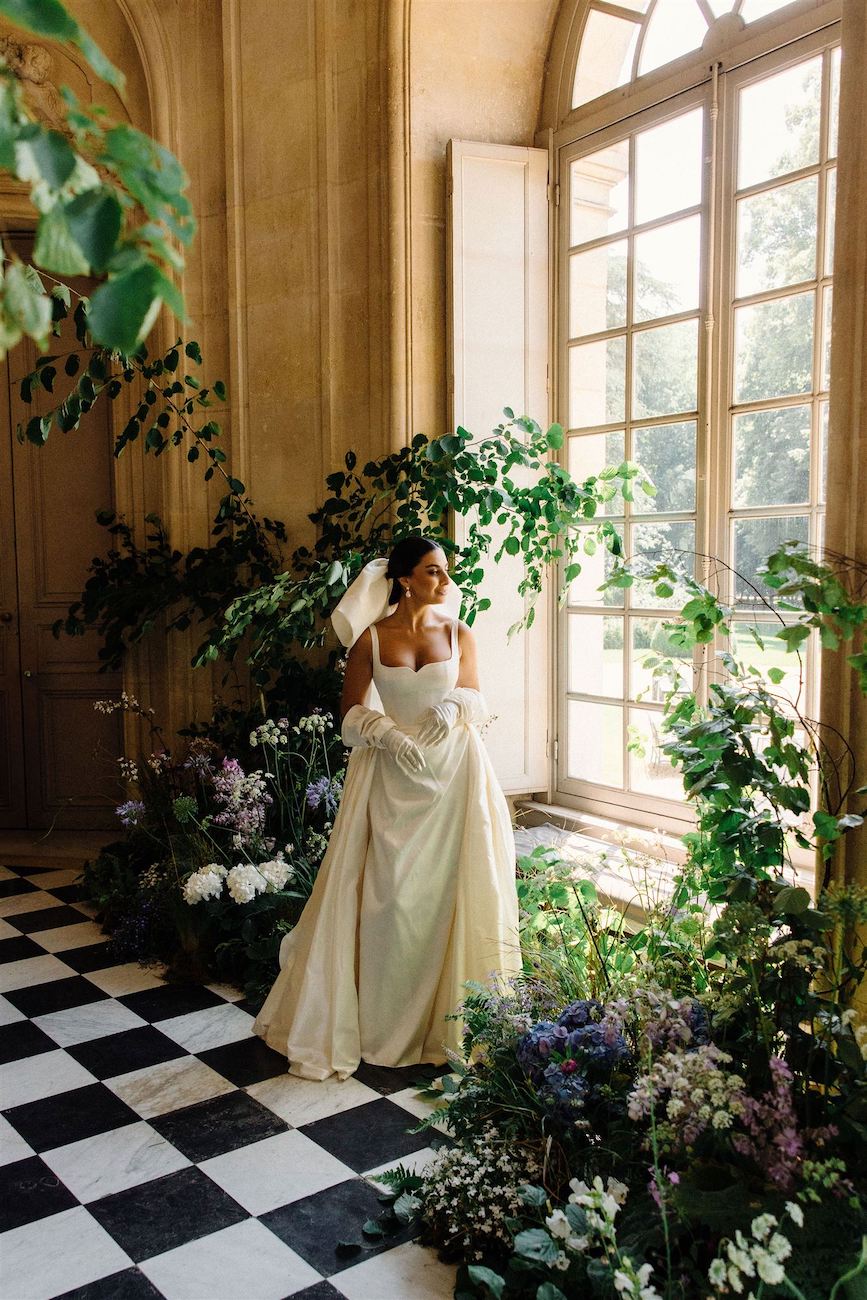 A woman in a white gown and gloves stands indoors on a black-and-white checkered floor, surrounded by green plants and flowers near large arched windows.