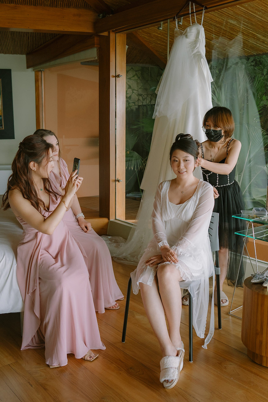 A bride sits on a chair while a hairstylist works on her hair. Two women in pink dresses sit nearby, with one taking a photo. A wedding dress hangs in the background.
