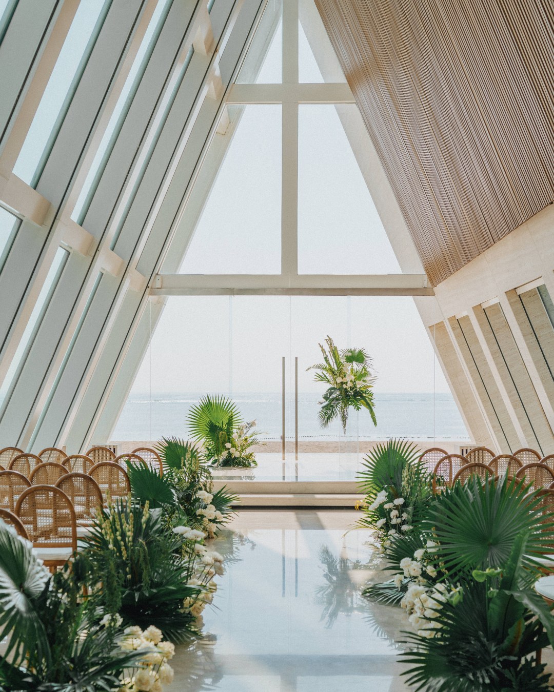 An airy, glass-walled chapel at Conrad Bali with wooden chairs and lush green floral arrangements facing large windows overlooking the ocean.