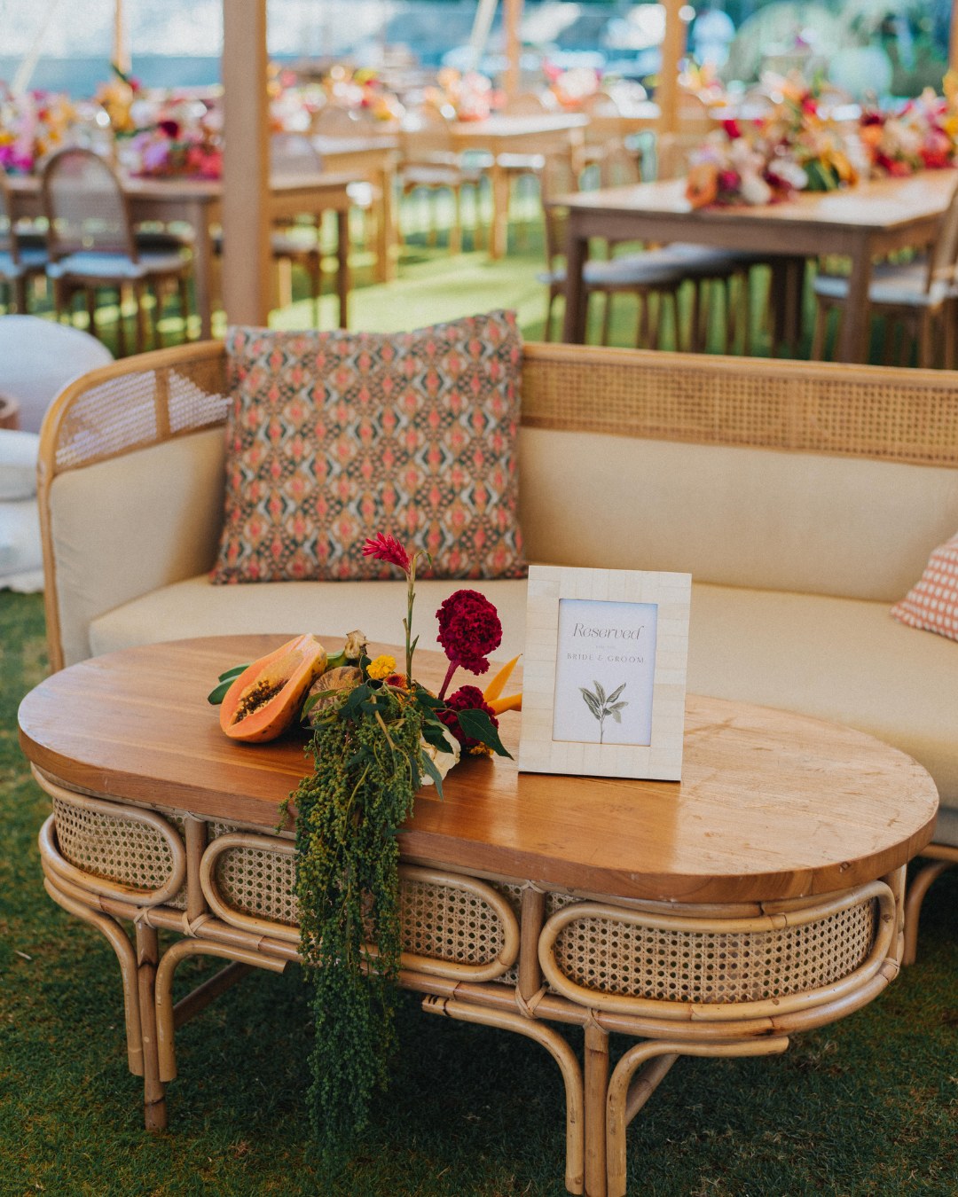 A wicker sofa with patterned cushions and a wooden coffee table decorated with flowers, greenery, and a framed card, set in an outdoor event space.