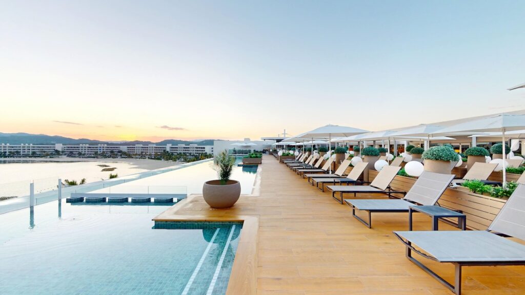 Rooftop infinity pool with lounge chairs, potted plants, and umbrellas at sunset, overlooking a cityscape.
