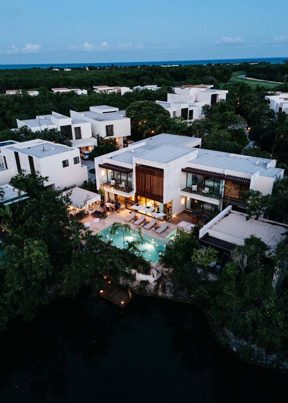 Aerial view of a modern, multi-story house with a lit swimming pool and lounge area, surrounded by trees and neighboring similar houses at dusk.