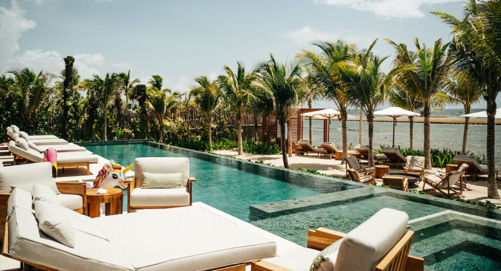 A luxurious outdoor pool area with lounge chairs, palm trees, and ocean views under a partly cloudy sky.