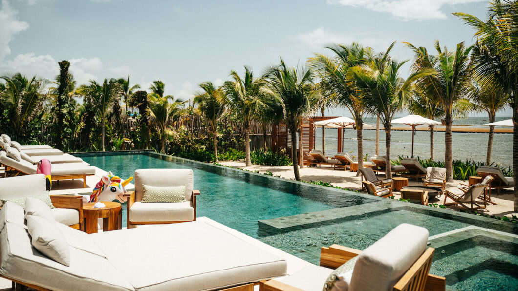 A luxurious outdoor pool area with lounge chairs, palm trees, and ocean views under a partly cloudy sky.