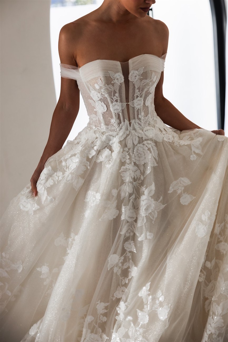 A woman wearing an off-the-shoulder, strapless white wedding gown with a sheer corset bodice and floral lace detailing.