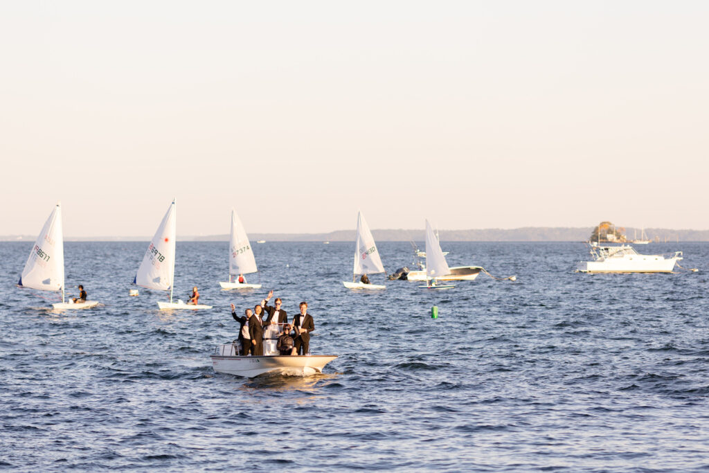 A group of people in suits stand on a small motorboat, waving, with several sailboats on the water in the background.
