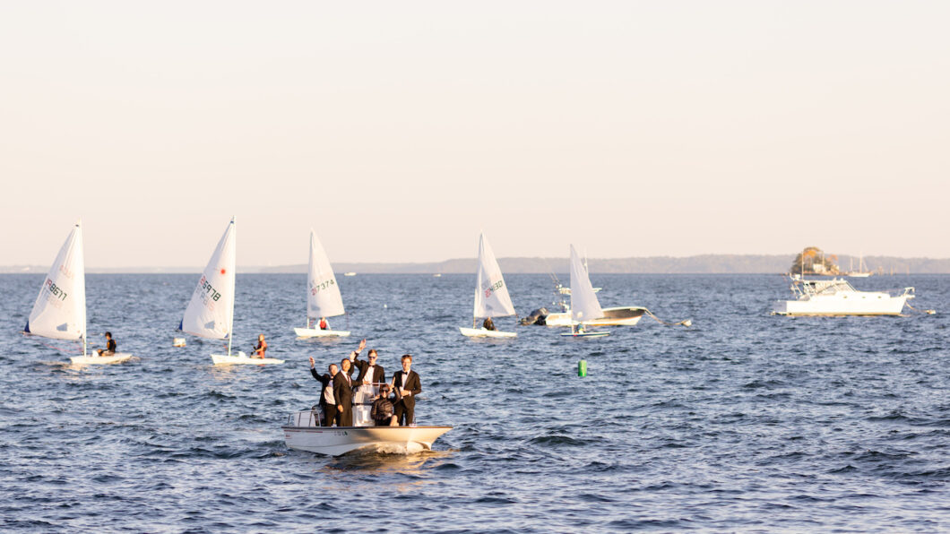 A group of people in suits stand on a small motorboat, waving, with several sailboats on the water in the background.