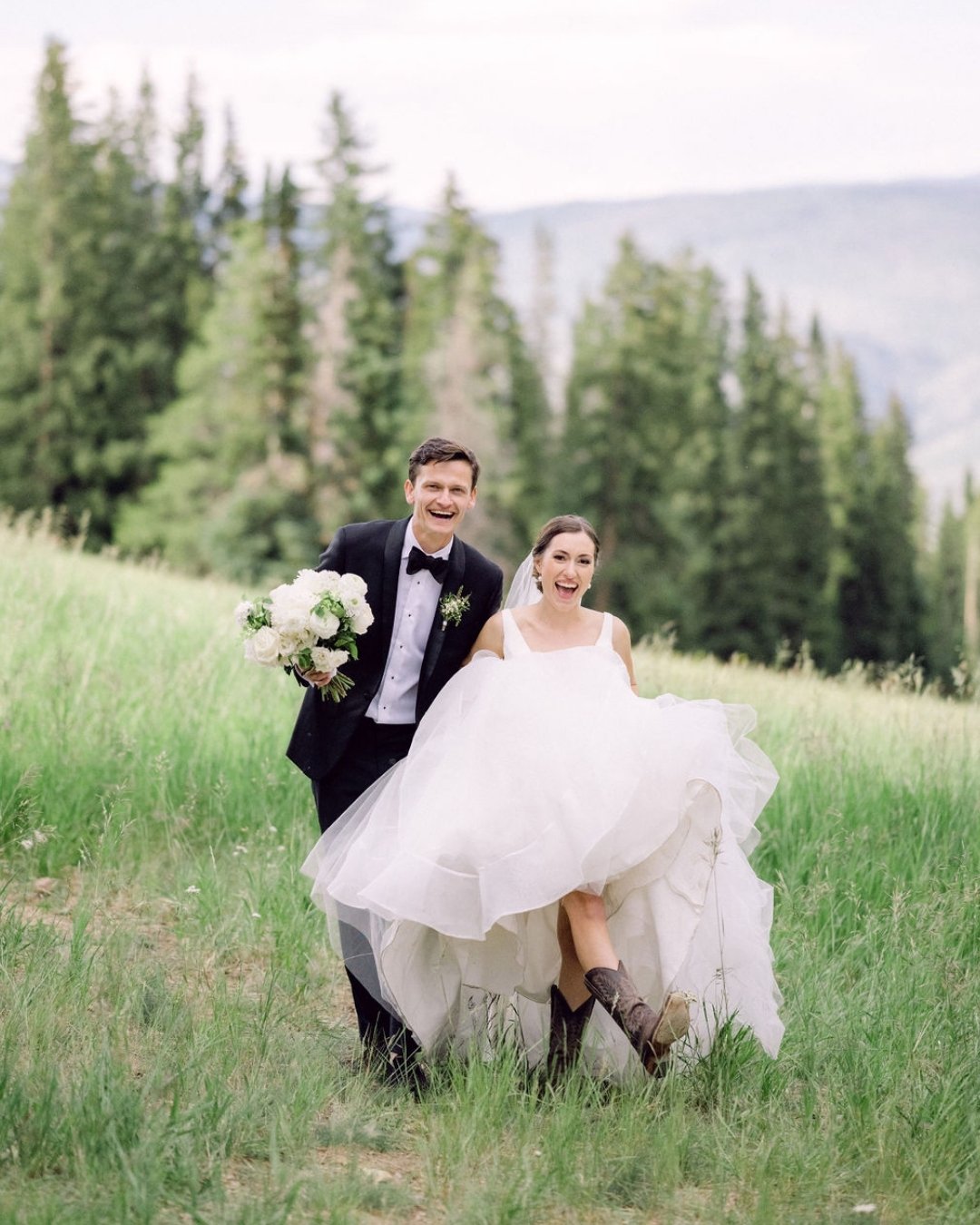 A bride in a white dress and boots and a groom in a black tuxedo walk together through a grassy field with trees in the background.