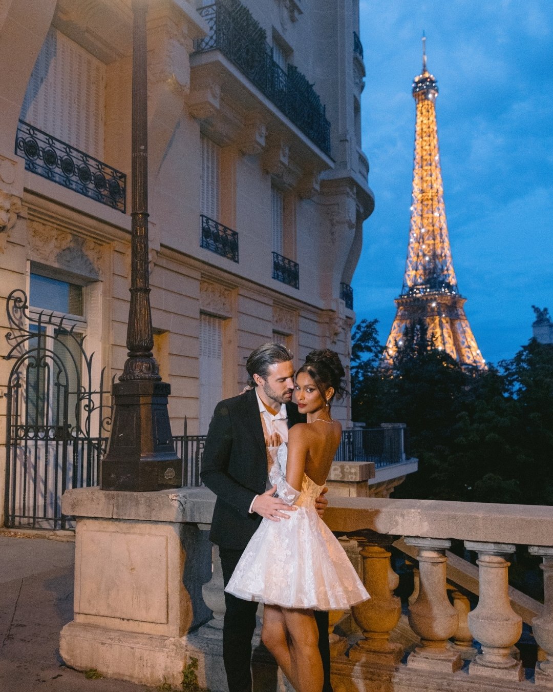 A couple dressed formally poses together on a Paris street at dusk, with the illuminated Eiffel Tower visible in the background.