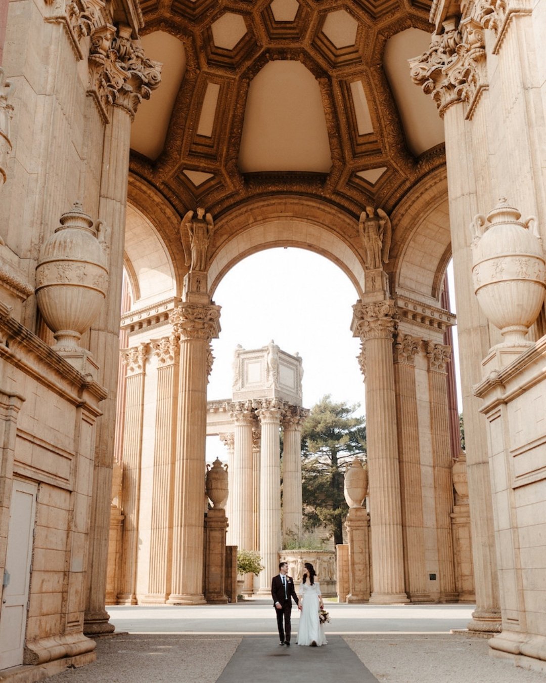 A couple in formal attire walks beneath large, ornate stone arches at an outdoor architectural site.