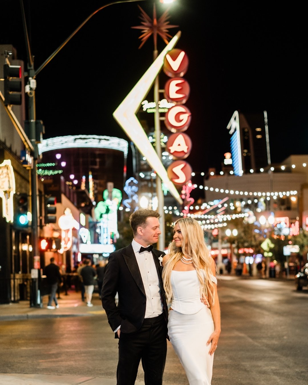 A couple dressed formally poses together on a street at night in downtown Las Vegas, with neon lights and a large "VEGAS" sign in the background.