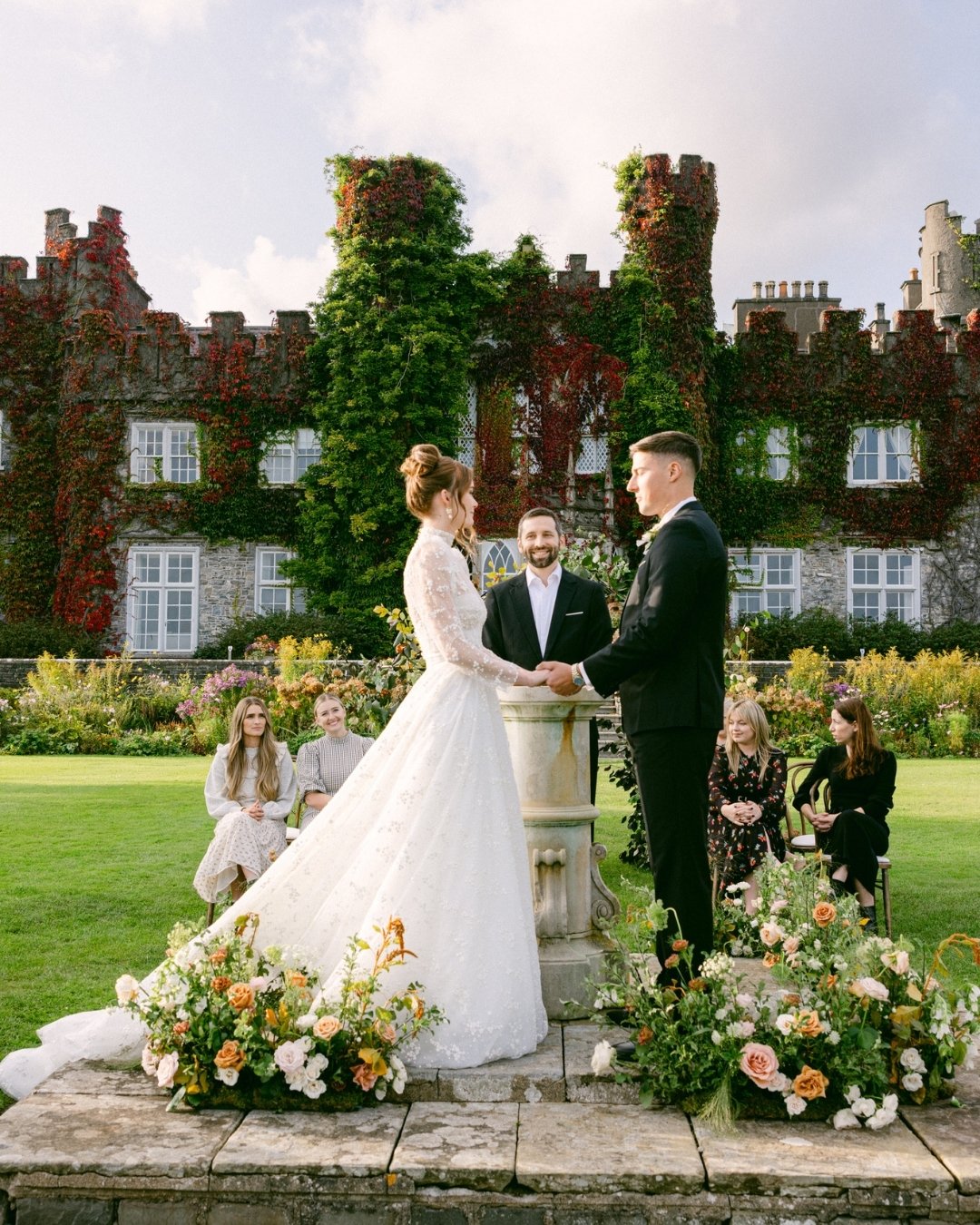A bride and groom stand holding hands at an outdoor wedding ceremony, with a historic ivy-covered building in the background and guests seated nearby.