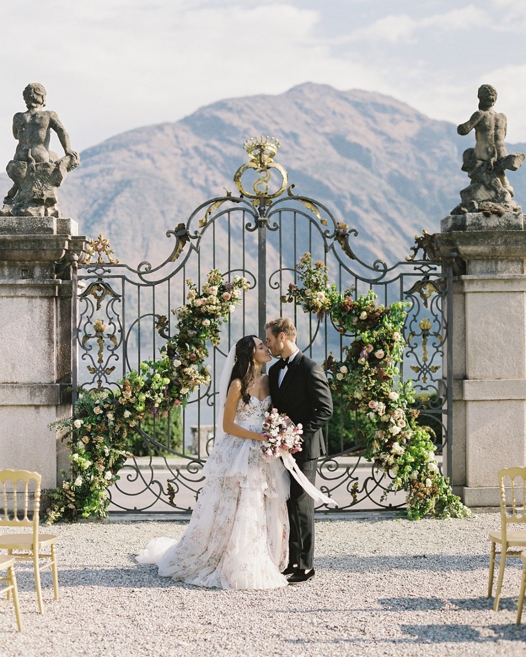 A bride and groom stand kissing in front of an ornate iron gate decorated with flowers, with mountains in the background.