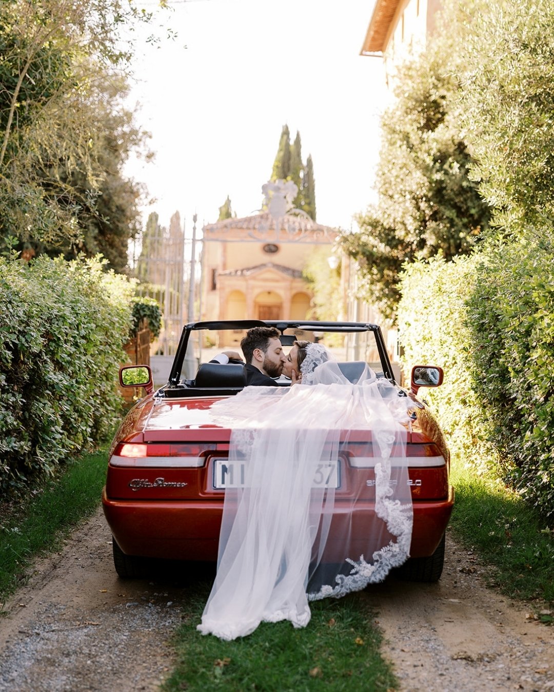 A bride and groom sit in a red convertible with the bride’s veil draped over the back, driving down a garden path toward an elegant building.