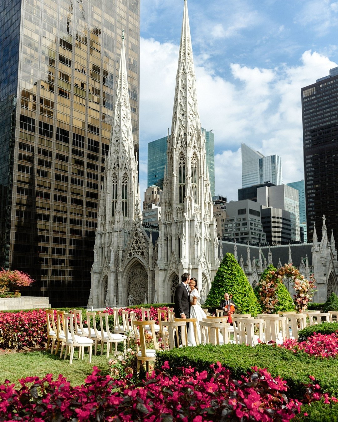 A couple stands on a rooftop garden decorated with flowers and chairs, with a gothic-style cathedral and modern skyscrapers in the background.