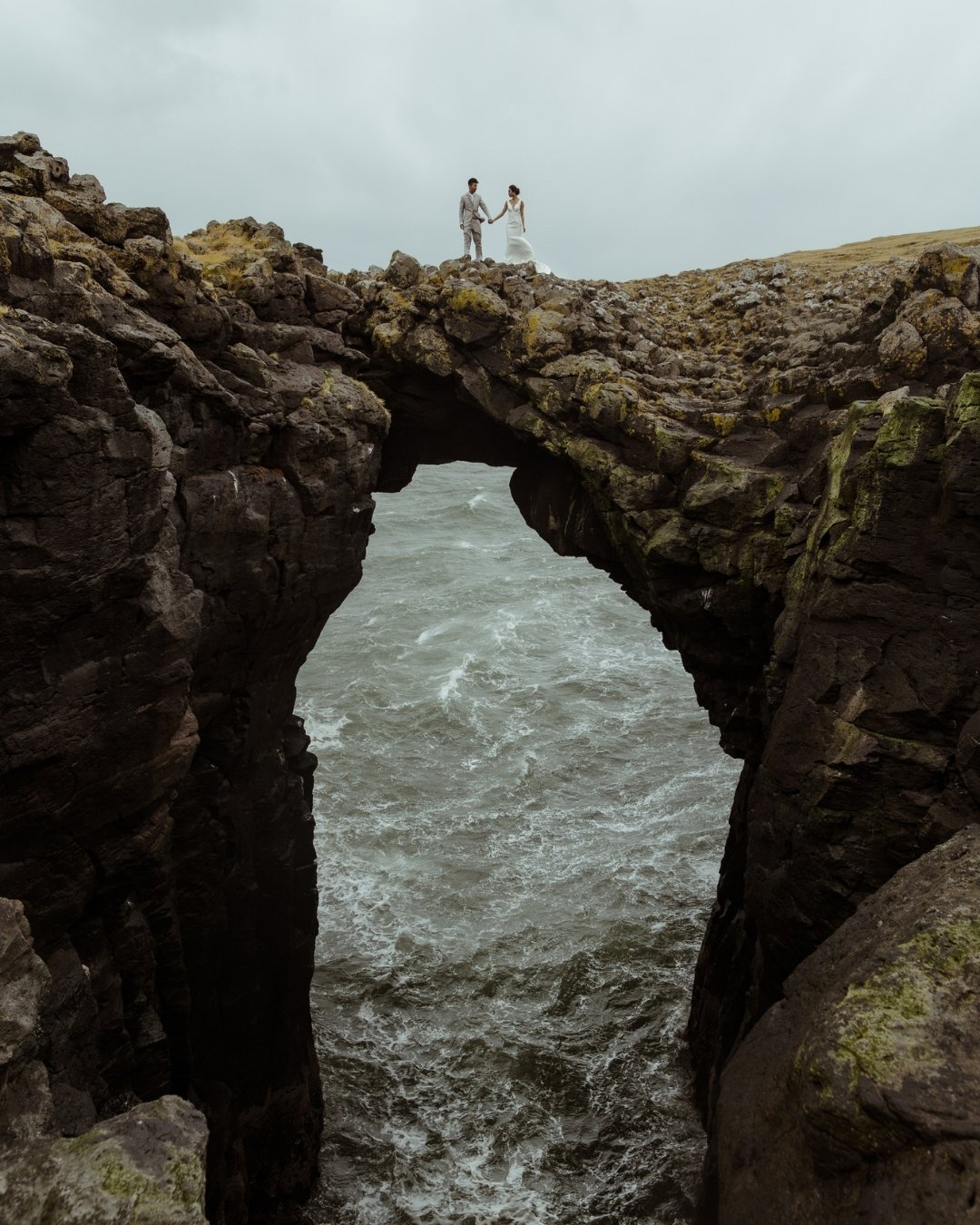 Two people stand on a natural rock arch above rough ocean waters, with an overcast sky in the background.
