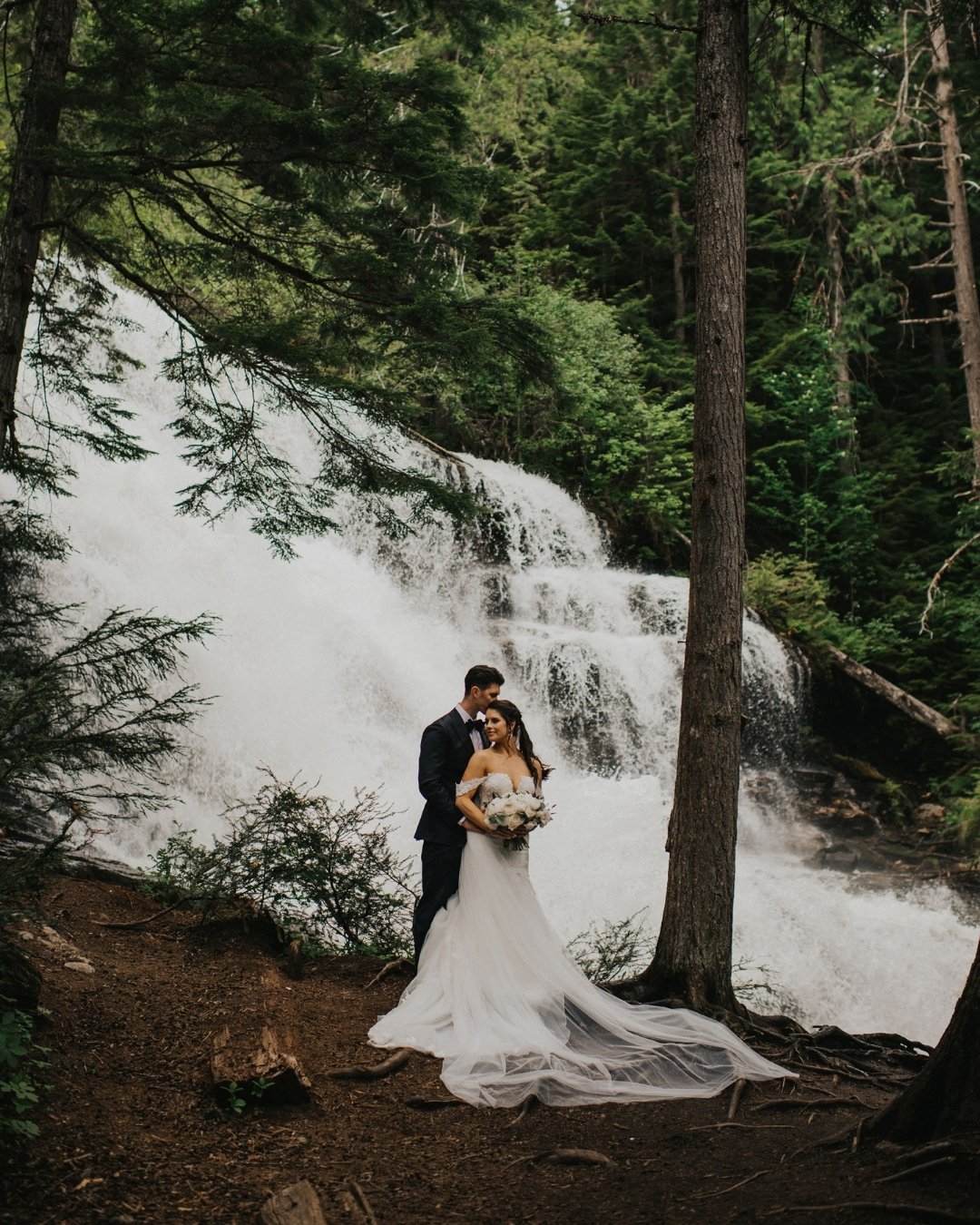A bride and groom stand together in a forest clearing in front of a large, cascading waterfall.