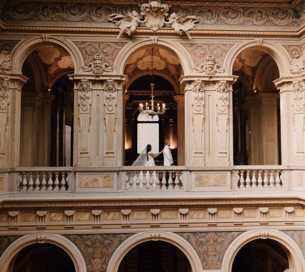 A couple dressed in wedding attire stands together on the balcony of an ornate, historic building with arched windows and detailed carvings.