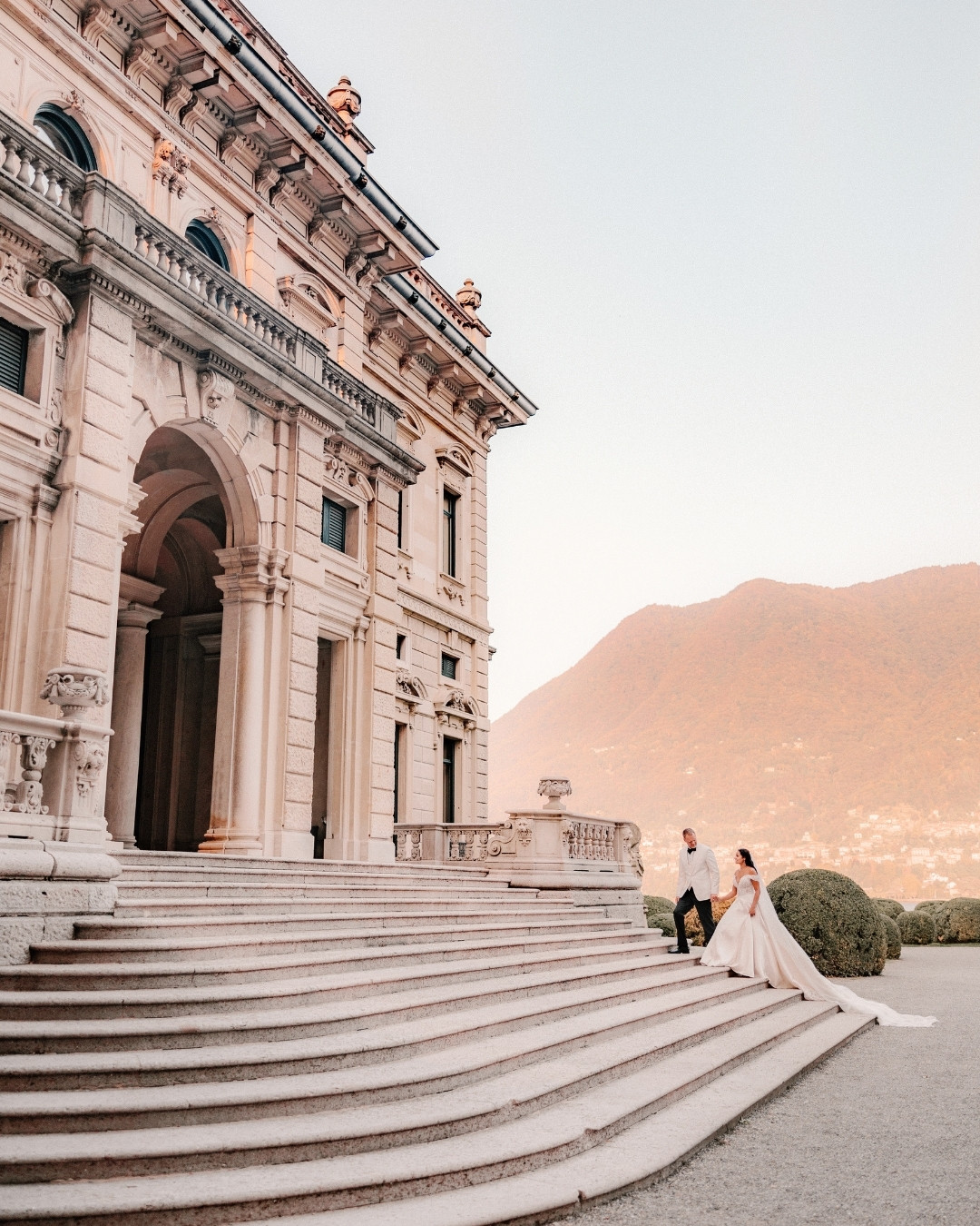 A bride and groom walk down the steps of an ornate, historic building with mountains and greenery in the background.