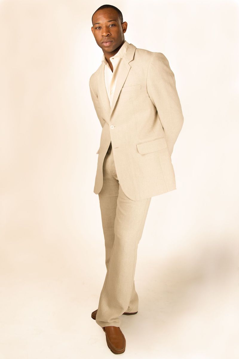 Man standing and posing in a beige suit with a white shirt and brown shoes against a plain light background.
