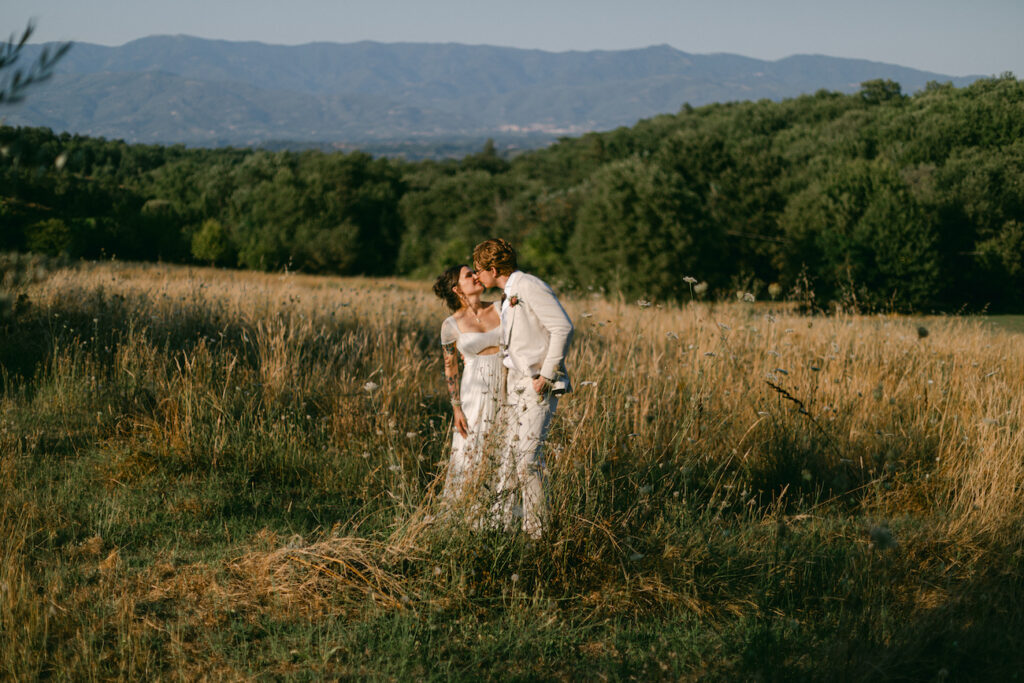 A couple dressed in white stands in a grassy field, sharing a kiss, with hills and trees in the background under a clear sky.