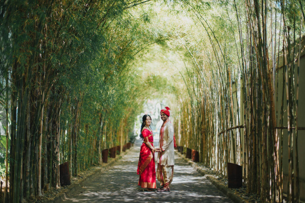 A couple in traditional Indian attire stands holding hands on a pathway beneath a green bamboo archway.