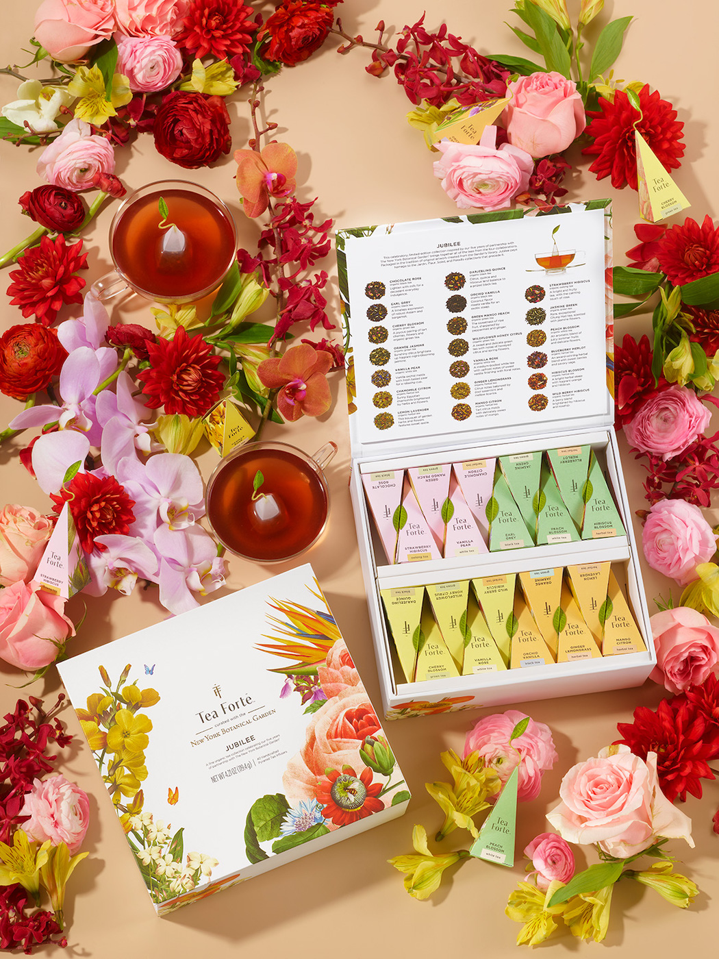 An open floral tea sampler box with assorted tea bags, a tea guide, two cups of tea, and various fresh flowers arranged on a beige surface.