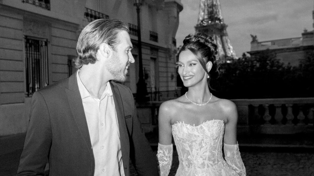 A couple dressed formally, with the woman in a wedding dress, smiles at each other while walking outdoors near the Eiffel Tower at night.