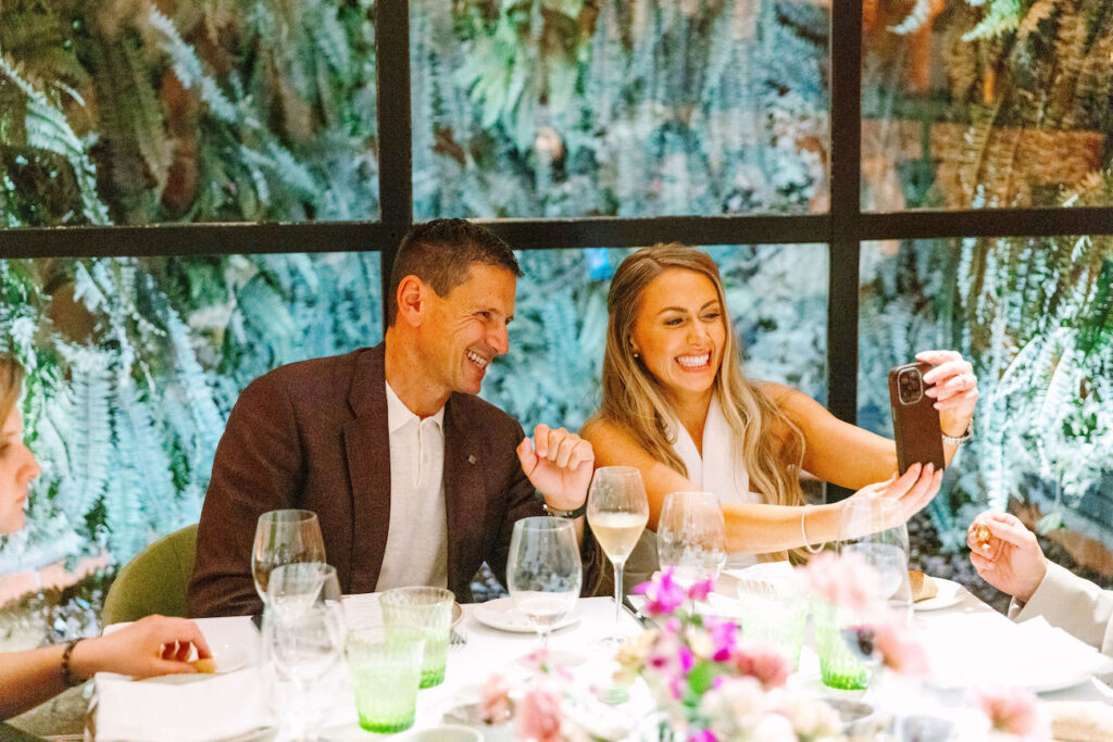 A man and woman sit at a table set for a meal, smiling and taking a selfie together, with glassware and floral decorations in front of them.