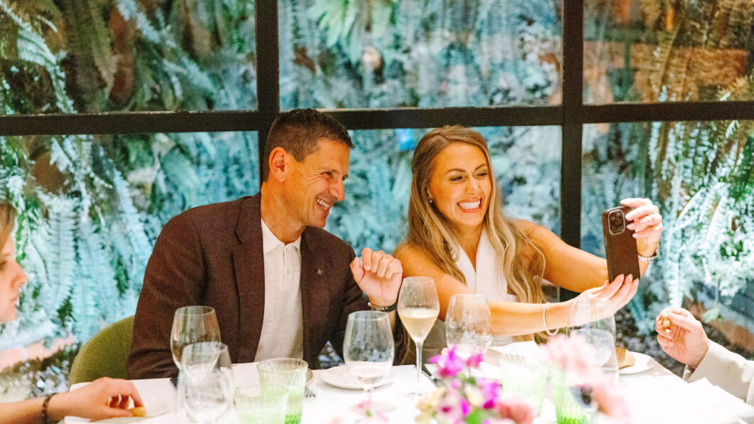 A man and woman sit at a table set for a meal, smiling and taking a selfie together, with glassware and floral decorations in front of them.