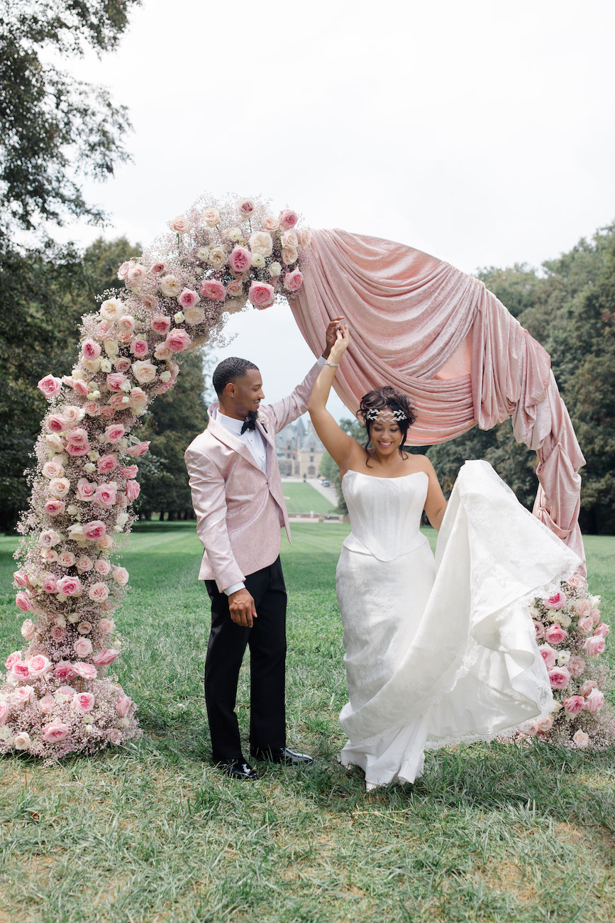 A bride and groom stand under a floral and fabric wedding arch outdoors. The groom holds up part of the arch as the bride lifts her dress and smiles.