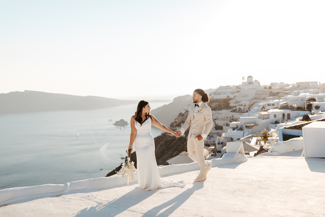 A bride and groom hold hands and walk on a white terrace overlooking the sea and white buildings, likely in Santorini, Greece, under bright sunlight.