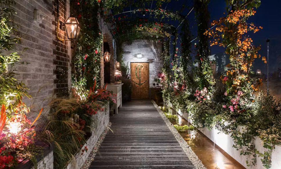 A wooden walkway with surrounding greenery, flowering plants, and lantern lights leads to a closed wooden door at night.