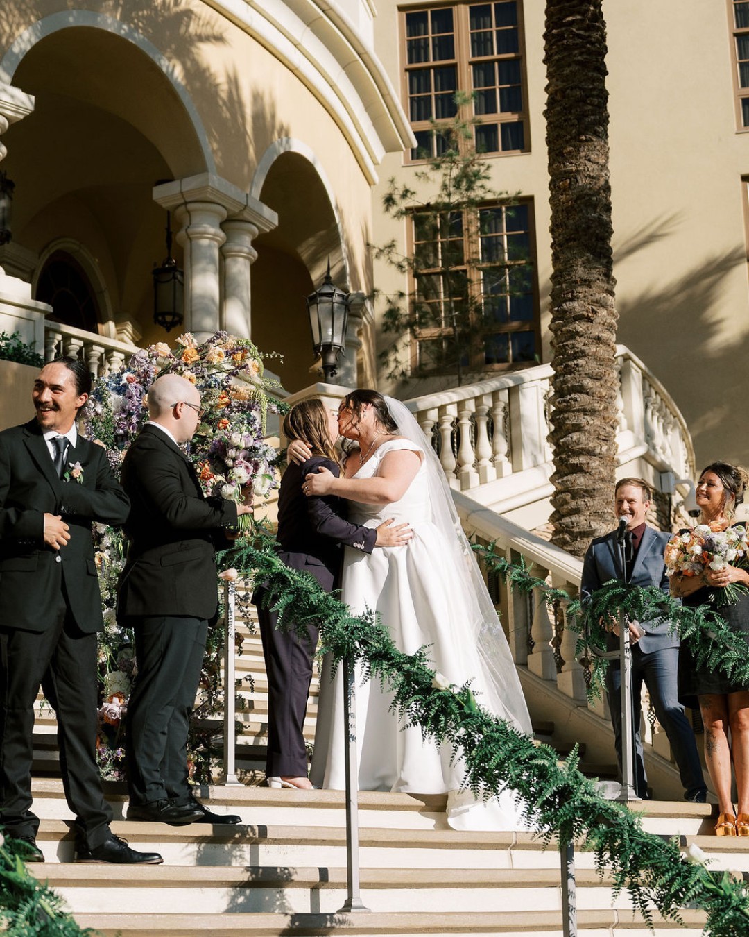 A bride in a white dress embraces and kisses a woman on the stairs of an outdoor venue, with wedding guests and floral decorations in the background.