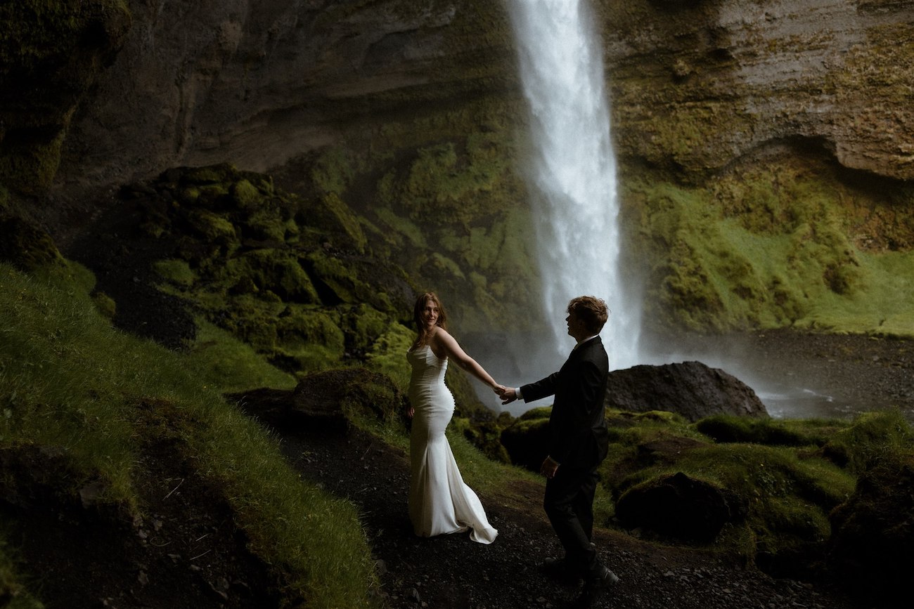 A bride and groom stand holding hands on a rocky path with lush greenery, near a tall waterfall cascading down a cliff.
