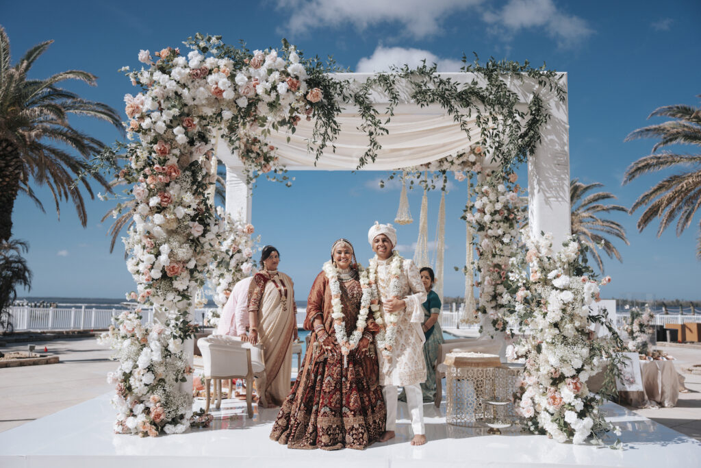 A bride and groom in traditional South Asian attire stand under a floral wedding canopy outdoors, accompanied by family, with palm trees and blue sky in the background.