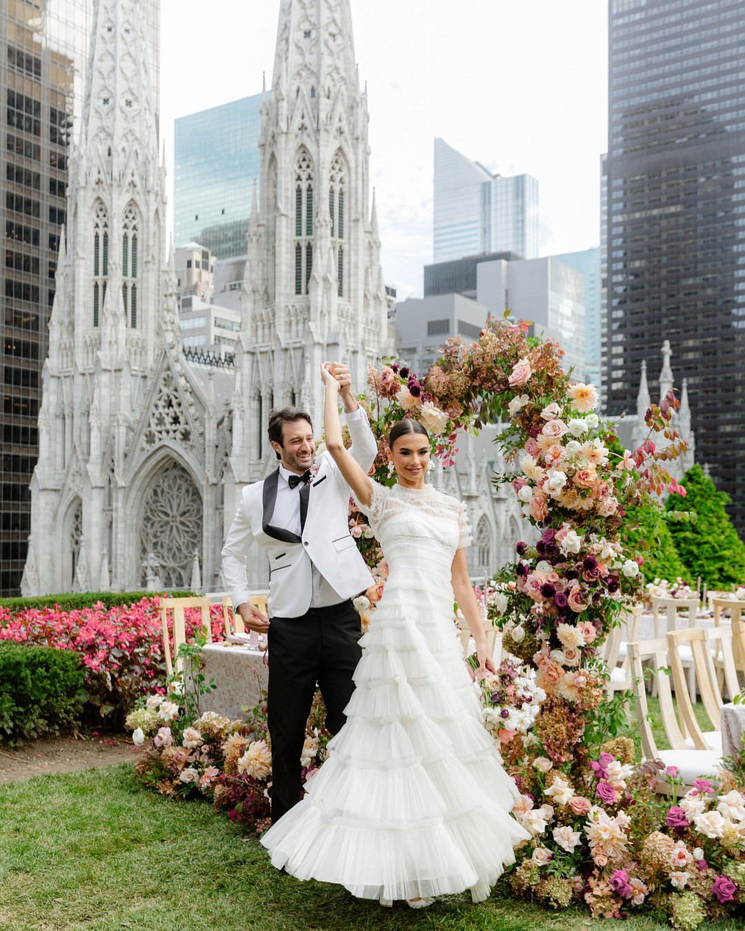A newlywed couple stands under a floral arch in formal attire, celebrating outside near a Gothic-style cathedral and modern skyscrapers.