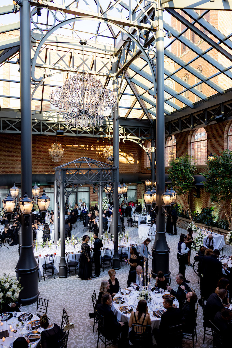 Guests seated at round tables in an elegant, glass-ceilinged venue with tall iron columns, chandeliers, and natural light during a formal event.