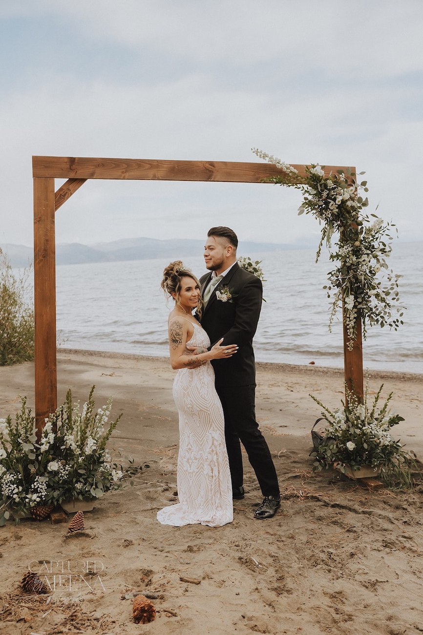 A bride and groom stand together under a wooden arch decorated with greenery and flowers on a sandy beach, with water and sky in the background.
