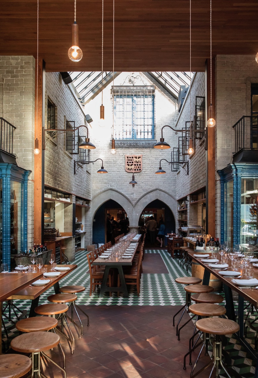 A spacious restaurant interior with long communal tables, wooden stools, high ceilings, hanging lights, and arched doorways at the far end.