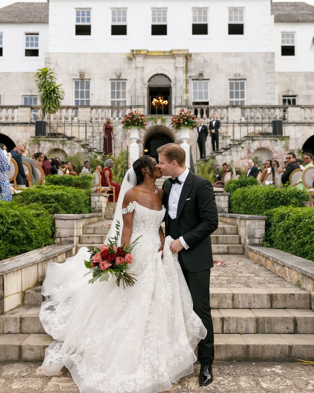 A bride and groom stand close together on stone steps outside a large white building, surrounded by guests and greenery.