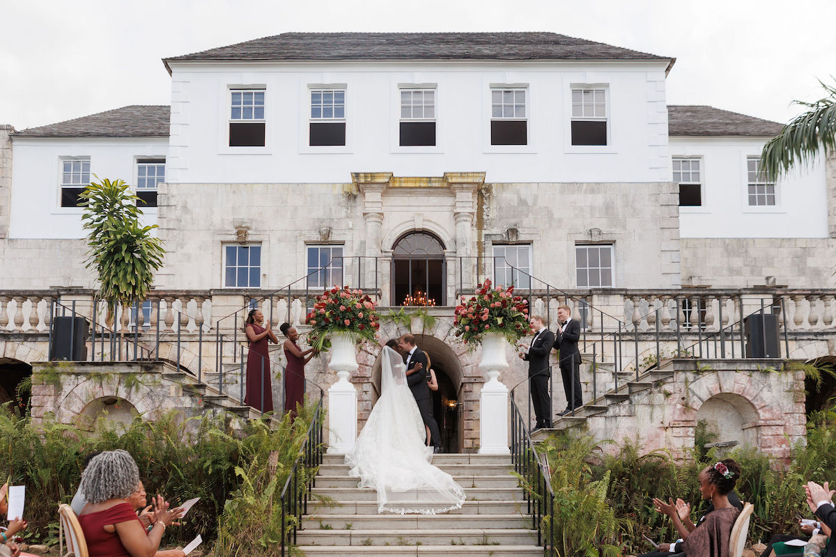 A bride and groom kiss on the steps of a large stone building, surrounded by their wedding party and guests.