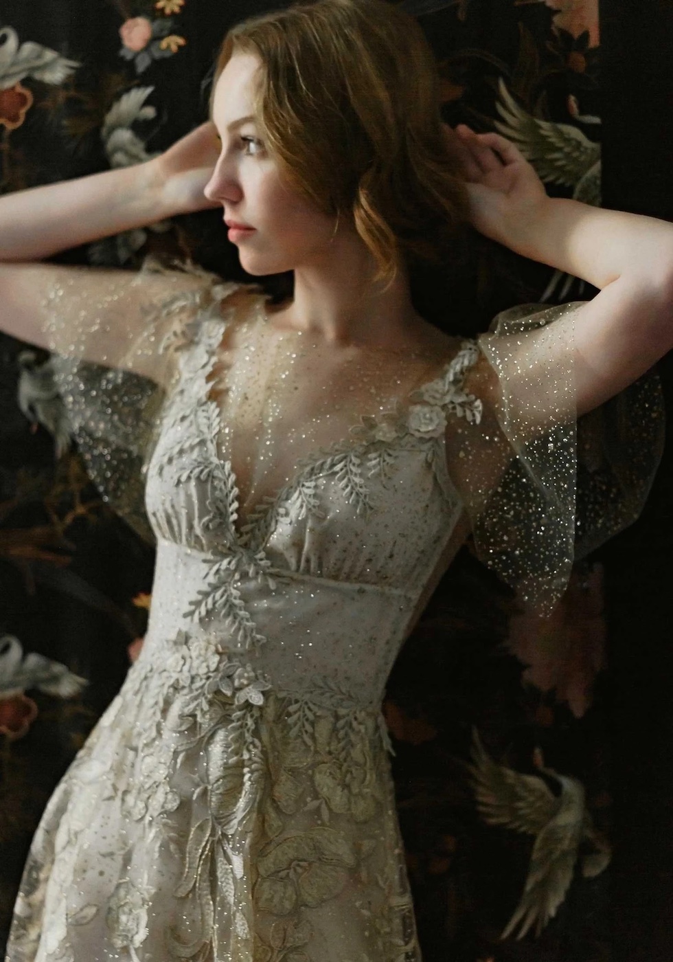 A woman in a light-colored, embroidered dress with sheer sleeves stands in front of a dark, floral-patterned background.