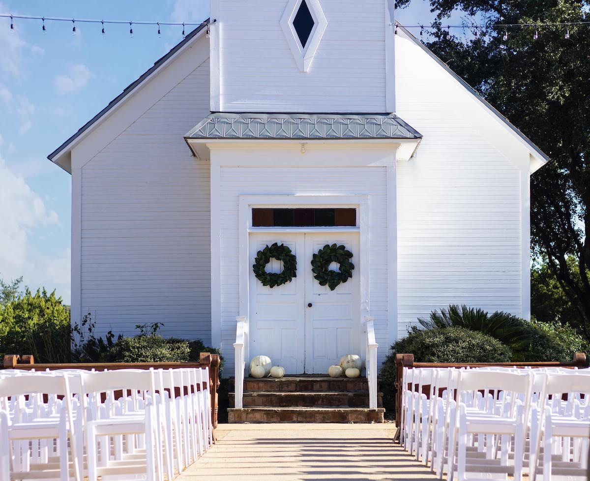 White chapel with two wreaths on the doors, white chairs arranged in rows on either side of an aisle, and trees in the background.