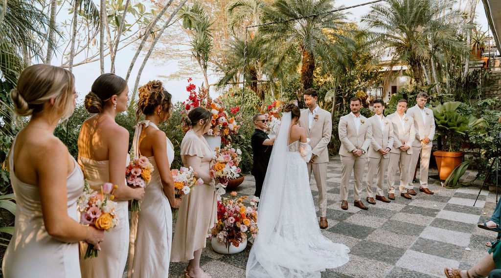 A bride and groom stand at the altar during an outdoor wedding ceremony, surrounded by bridesmaids and groomsmen in matching beige attire, with tropical plants in the background.