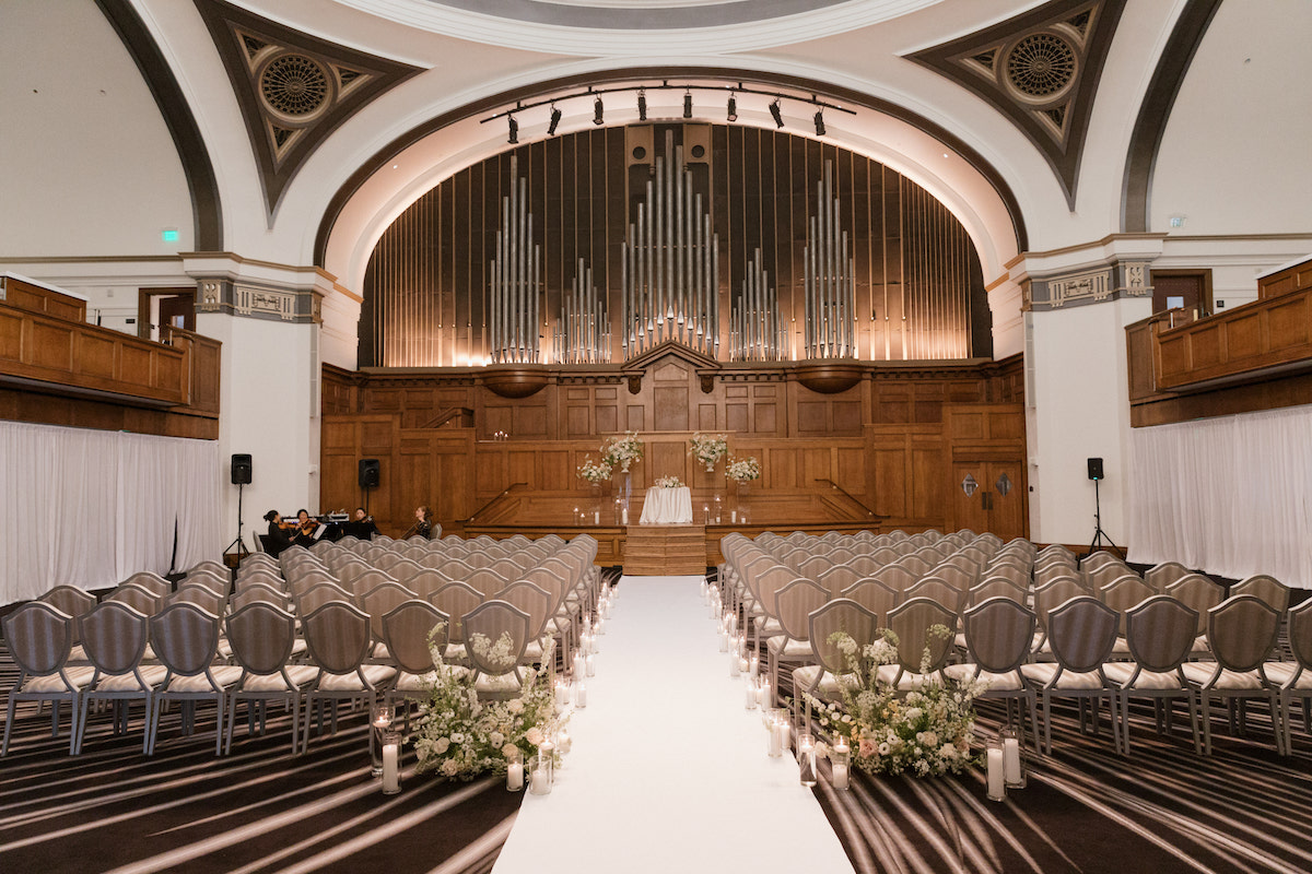 A formal indoor wedding venue with rows of chairs facing an altar, organ pipes in the background, and floral arrangements lining the aisle.