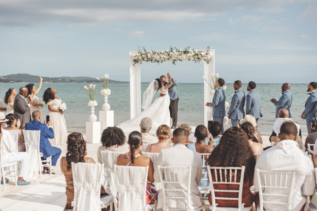 A bride and groom kiss under a floral arch on a beach as wedding guests seated outdoors look on.